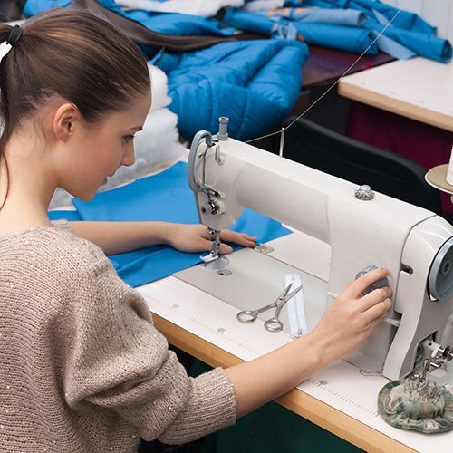 Eine junge Frau mit langen braunen Haaren sitzt an einer N&auml;hmaschine und stellt an dieser etwas ein. Ein blauer Stoff liegt zum N&auml;hen bereit