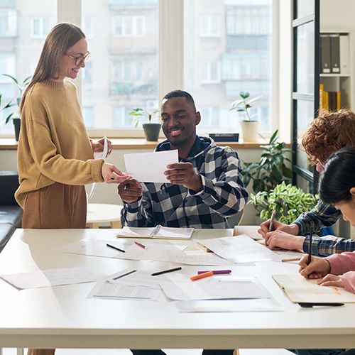 An einem eckigen wei&szlig;en Tisch sitzen drei Teilnehmende. Eine Dozentin steht am Tisch und zeigt, ein junger Mann of Colour zeigt ihr ein Blatt Papier. Die anderen beiden notieren etwas in ihre Unterlagen. Im Hintergrund sind Pflanzen und ein gro&szlig;es Fenster zu sehen.