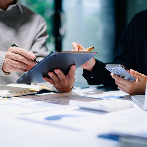 Nahaufnahme eines Tischs, an dem ein Mann ein Tablet h&auml;lt und eine Frau ein Handy. Sie zeigen sich gegenseitig etwas auf dem Tablet. Die Gesichter sind nicht zu sehen.