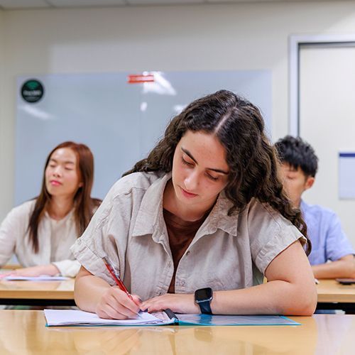 Mehrere Persone sitzen in einem Seminarraum und schreiben etwas auf und h&ouml;ren zu. Im Vordergrund ist eine Frau mit langen braunen Haaren, die etwas in ihr Heft notiert