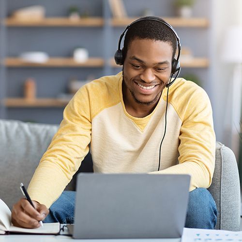 Ein Man of Colour sitzt auf dem Sofa, hat ein Headset auf und schaut l&auml;chelnd auf ein Laptop. Dabei notiert er etwas in ein Notizbuch.