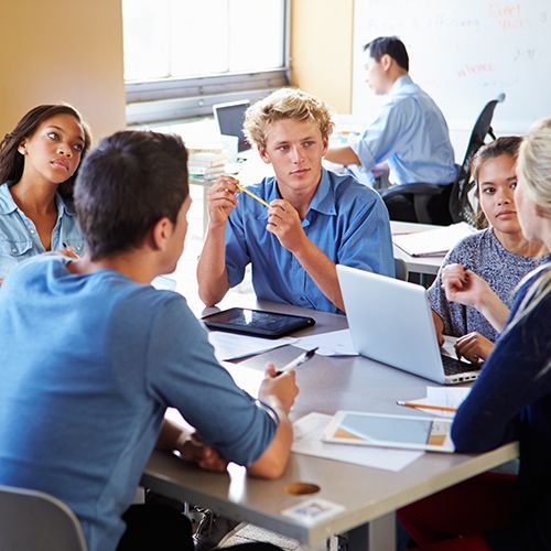 Mehrere junge Menschen unterschiedler Herkunft sitzen an einem Tisch zusammen. Sie haben Unterlagen, Laptop und Tablet auf dem Tisch und schauen auf eine junge Frau die man nur von hinten sieht.