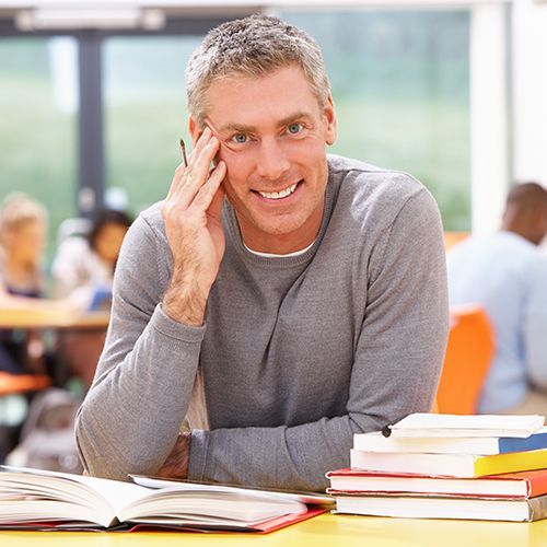Das Foto zeigt einen Mann in einem modernen Klassenraum. Er sitzt an einem Tisch mit B&uuml;chern und lacht in die Kamera.