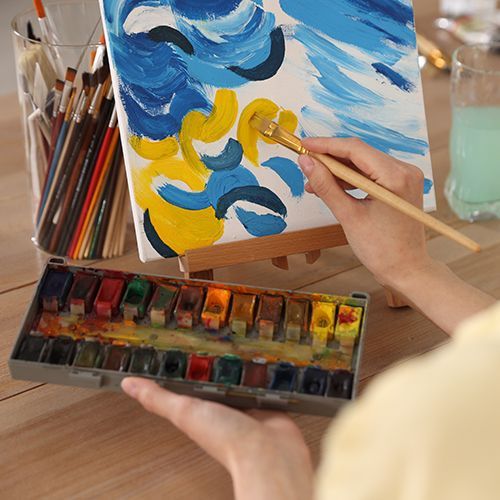 Auf einem Tisch steht eine kleine Staffelei mit Leinwand auf der mit blau, schwarz und gelb halbrunde Pinselstriche aufgetragen wurden. Ein Glas mit Pinseln steht dahinter. Eine Frau im Anschnitt ist mit Pinsel und Aquarellkasten dabei, weiter Farbe aufzutragen.