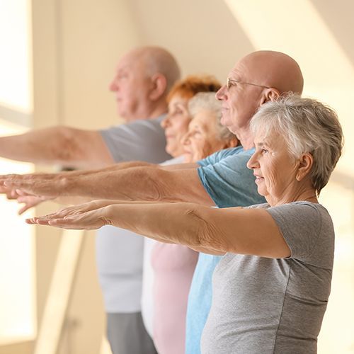 In einem hellen Sportraum stehen f&uuml;nf &auml;ltere Menschen seitlich hintereinander. Sie strecken ihre Arme und H&auml;nde nach vorne. Im Fokus ist eine Frau mit kurzen wei&szlig;en Haaren und ein Mann mit Brille und Glatze.