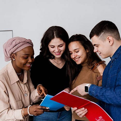 Eine Gruppe junger People of Colour stehen in einem Raum und schauen gemeinsam in ein blaues und ein rotes Heft. Sie zeigen sich dabei etwas. Alle l&auml;cheln.