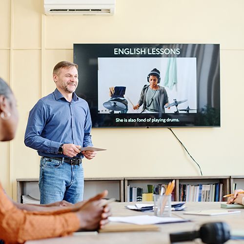 Ein Dozent steht in einem Seminarraum, hinter ihm ein Monitor mit einer Englisch-Lesson. Angeschnitten im Vordergrund einige Personne die zuh&ouml;ren.