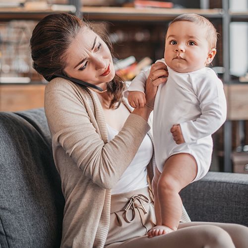 Eine Frau sitzt zu Hause auf dem Sofa. Sie hat das Telefon zwischen Kopf und Schulter geklemmt und kleidet nebenbei ihr Baby an.