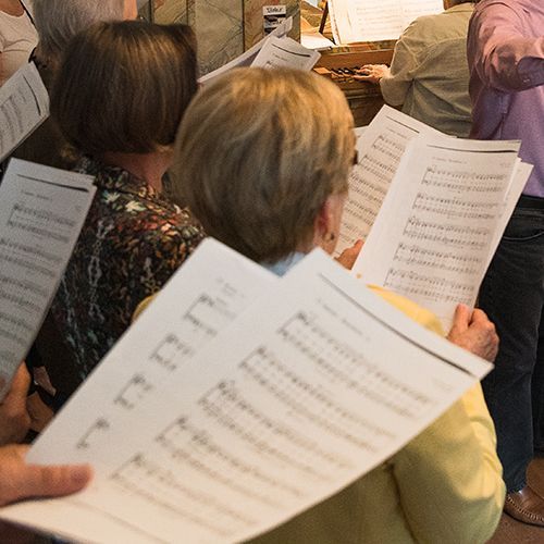 Menschen halten Notenbl&auml;tter und singen in einem Chor. 
In der Mitte steht der Dirigent