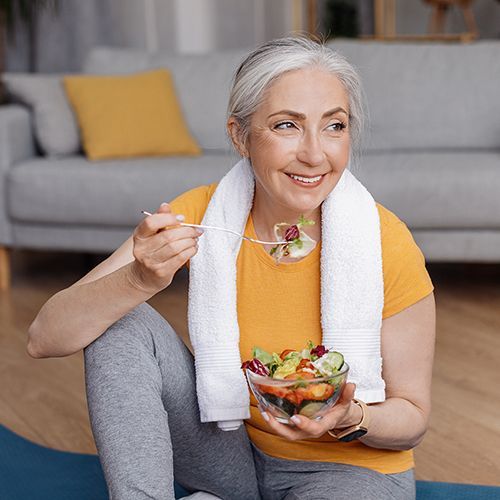 Eine &auml;ltere Frau sitzt mit einer Matte in einem Wohnzimmer vor einem Sofa und i&szlig;t aus einer gl&auml;sernen Sch&uuml;ssel Salat.