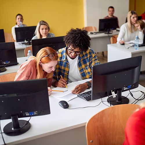 Mehrere Teilnehmer sitzen in einem Computerraum an Tischen, einzeln oder zu zweit. Im Fokus ist ein Mann mit Afro- und eine Frau mit roten Haaren. Sie schauen sich gemeinsam etwas in einem Heft an und zeigen mit Bleistiften darauf.