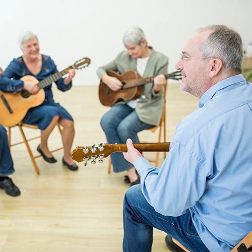 Eine Gruppe älterer Menschen sitzen auf Klappstühlen in einem hellen großen Raum. Sie haben Gitarren in der Hand. Sie schauen auf einen Mann, der im Fokus im Vordergrund von seitlich, hinten zu sehen ist.