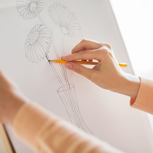 Eine Hand zeichnet auf ein weißes Papier, das auf einem Brett von einer Frau gehalten wird mit Bleistift drei Blumen in einer Vase.