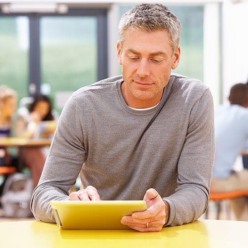 Ein Mann sitzt mit einem Tablet in einem Seminarraum und liest oder notiert etwas. Im Hintergrund sind weitere Personen unscharf zu erkennen.