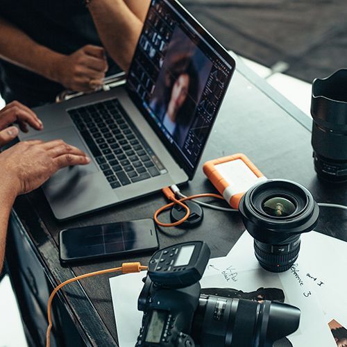 Zwei Personen arbeiten an einem Bild auf einem Laptop, daneben liegen Kameraequipment, Blätter und eine externe Festplatte.