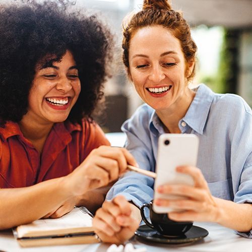 Zwei Frauen schauen lachend auf ein Smartphone und eine zeigt mit einem Stift darrauf.