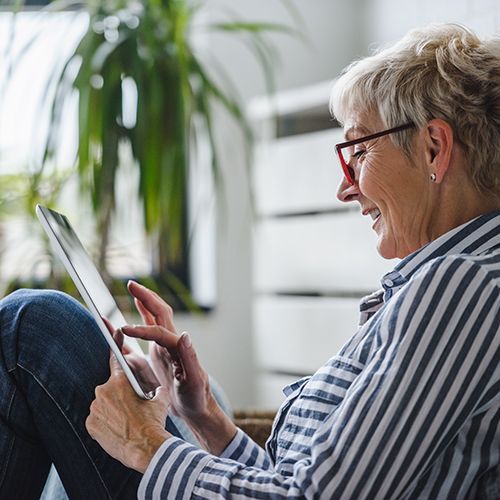 Eine ältere Frau sitzt mit dem Rücken an einer Kommode und hat ein Tablet in der Hand. Sie trägt ein gestreiftes Hemd und eine Brille. Sie lächelt.
Im Hintergrund sind Pflanzen unschwarf zu sehen.