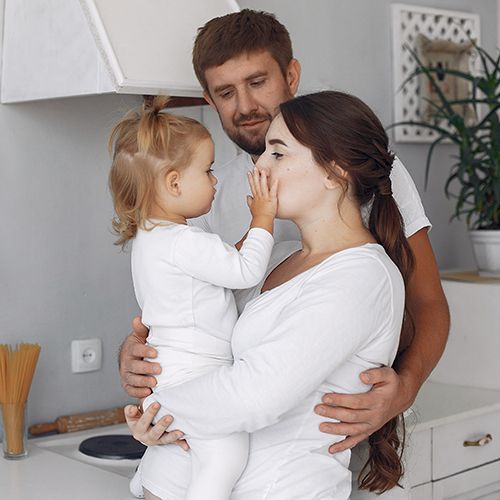 Eine Familit mit einer Tochter in ihrer Küche. Die schwangere Mutter hat die kleine Tochter auf dem Arm und der Vater steht dahinter. Mutter und Tochter schauen sich an.