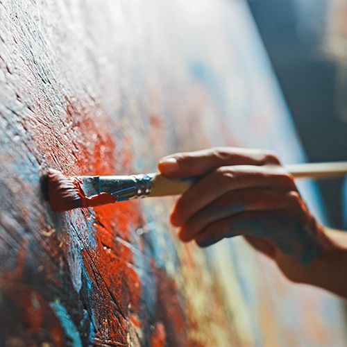 Eine Nahaufnahme eines Pinsels auf einer Leinwand mit roter Farbe, der von einer Frau gehalten wird, die man sehr unscharf im Hintergrund sieht.