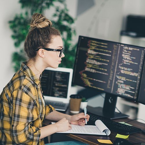 Eine junge Frau sitzt vor mehreren Monitoren mit Programmcode und notiert etwas auf Papier, das sie vor sich in einem Klemmbrett liegen hat.
