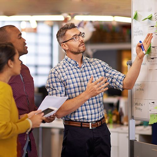 An einem Whiteboard stehen drei Personen und betrachten daran aufgehängte Zettel.