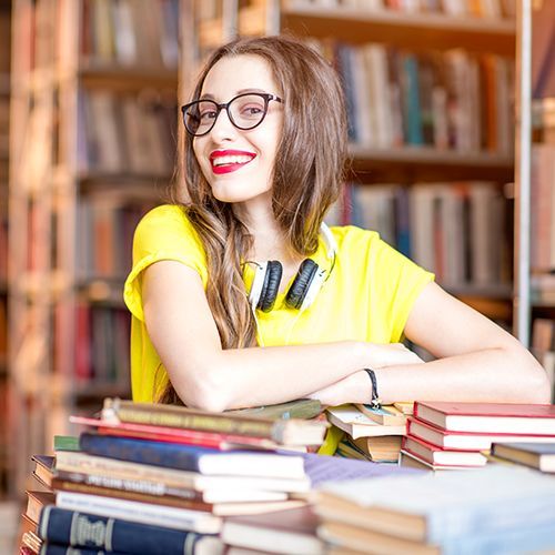 Eine Studentin sitzt in einer Bibliothek. Sie trägt Kopfhörer um ihren Hals und vor ihr liegen auf einem Tisch verstreut viele unterschiedliche Bücher. Sie lächelt stolz in die Kamera.