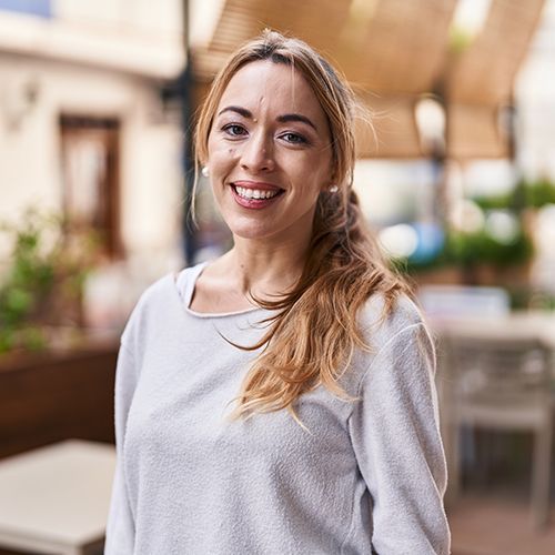 Eine Frau schaut lächelnd in die Kamera. Sie hat einen hellen Pullover an und im Hintergrund sind unscharf Pflantzen und Outdoor-Möbel zu sehen.