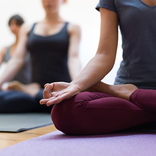 Auf einem Holzboden liegen Yogamatten auf denen Frauen im Lotussitz schräg hintereinander sitzen. Sie tragen Sportkleidung und halten die Hände in einer Meditationspose auf den Knien. Bei der vorderste Frau im Fokus ist das Gesicht nicht zu sehen. Die hinteren Frauen sind unscharf