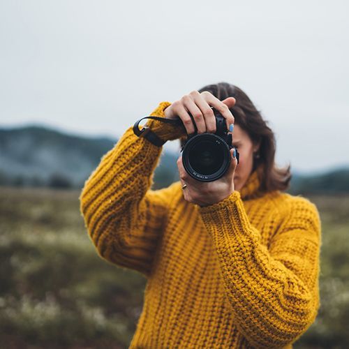 Eine Frau mit gelbem Strickpullover hält eine digitale Spiegelreflexkamera vor ihr Gesicht. Sie steht in der Natur.