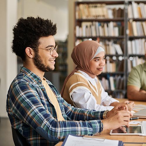 Drei People of Colour sitzen an einem Tisch. Vor ihnen liegen Papier, Laptops und Stifte. Ein Mann im Fokus schaut im Profil rechts aus dem Bild heraus.