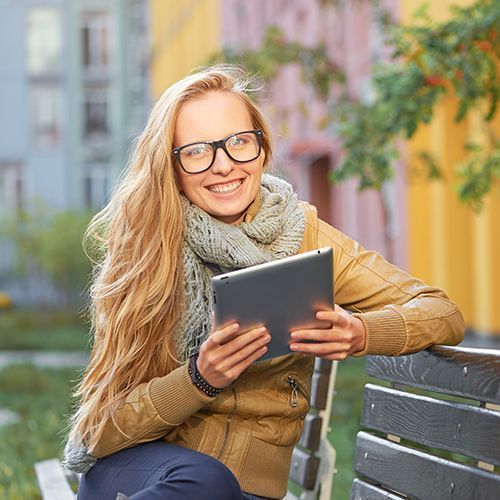 Eine junge Frau mit Brille sitzt auf einer Bank und hat ein Tablet in der Hand. Sie ist herbstlich angezogen.
