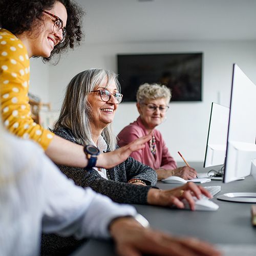 An einem langen grauen Tisch sitzen mehrere Personen an beiden Seiten. Sie schauen auf Monitore oder Laptops. Eine Person steht auf der linken Seite und zeigt einer Teilnehmerin etwas auf dem Monitor.