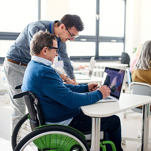 In einem Kursraum sitzen Teilnehmer an dreieckigen Einzeltischen mit Laptops vor sich. Im Fokus ist ein Mann im Rollstohl über den sich dahinter jemand zum Bildschirm des Laptsops beugt, Im Hintergrund sind große Fenster unscharf zu erkennen.