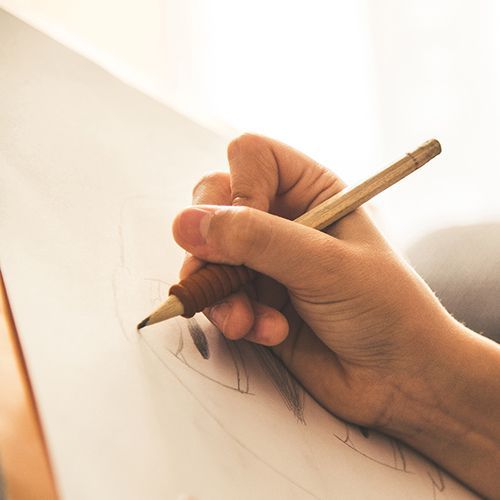 Eine Hand hält einen Bleistift und zeichnet auf ein Blatt Papier, das auf einem Holzbrett liegt.