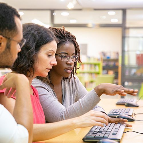 Drei People of Colour sitzen an Computern an der Wand. Die Frau in der Mitte tippt, die rechts neben ihr zeigt etwas auf dem Bildschirm. Der Mann links hört zu.