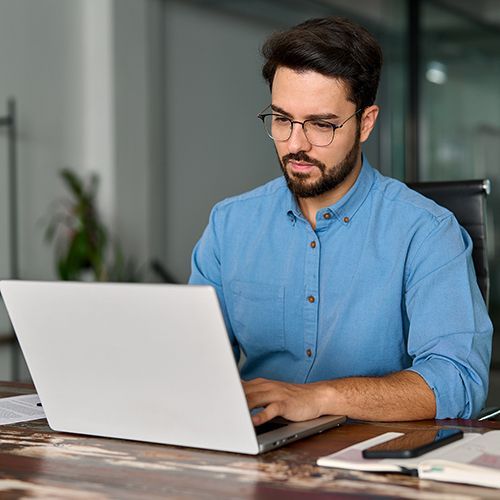 Ein junger Mann sitzt in einem Raum mit Glaswänden vor einem Laptop auf einem Holztisch und schaut konzentriert.