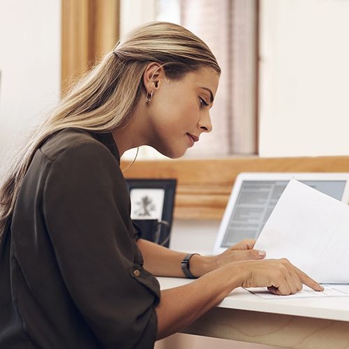Eine junge Frau sitzt an einem Tisch an einer Wand und blättert durch Unterlagen. Neben ihr steht ein Laptop, an der Wand hängt ein großer Spiegel im Holzrahmen