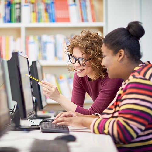 Lehrkraft erklärt einer Teilnehmenden eines Computer-Kurses etwas am Computer mit einem Bleistift in der Hand.