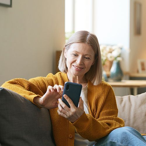 Eine ältere Frau sitzt seitlich auf einem Sofa und tippt etwas in ein Smartphone. Im Hintergrund ist das warme Licht einer Lampe.