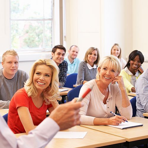 Das Foto zeigt eine Klassenraum-Situation mit erwachsenen Kursteilnehmer:innen. Sie sehen ihren Dozenten lächelnd an.
