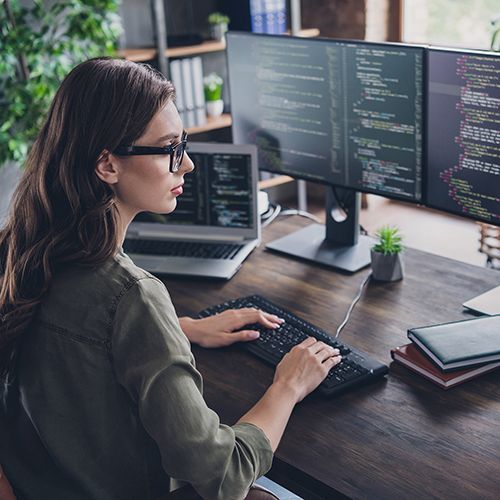 Eine Frau sitzt vor mehreren Bildschirmen mit Code-Zeilen. Neben ihr steht je ein Laptop und eine weitere Tastatur liegt vor ihr.