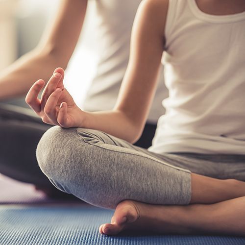 Auf Sportmatten sitzen zwei Frauen hintereinander im Lotussitz. Man sieht ihre Gesichter nicht. Sie tragen wei&szlig;-graue Sportkleidung.