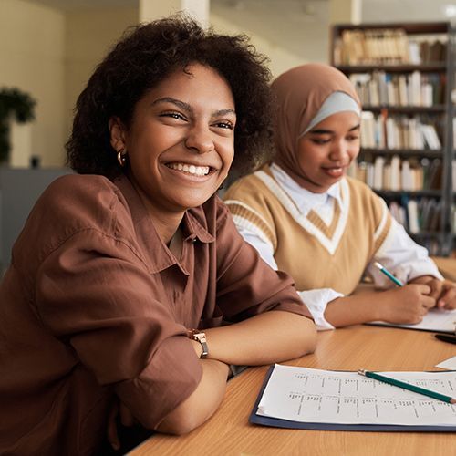 Drei Menschen of Colour sitzen an einem Tisch voller Papier, einem Laptop, einem Klemmbrett und Stiften. Im Hintergrund ist ein B&uuml;cherregal zu sehen. Im Fokus ist eine junge Frau die lachend an der Kamera rechts vorbeischaut.