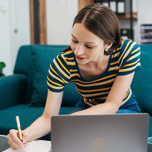 Eine Frau sitzt vornübergebeugt auf einem Sofa und hat auf dem Tisch vor ihr einen Laptop. Sie notiert etwas auf einem Block.