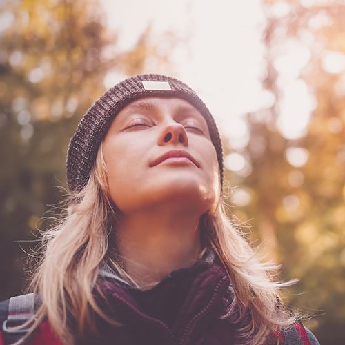 Eine Frau in Nahaufnahme. Sie tr&auml;gt eine M&uuml;tze, hat die Augen geschlossen und reckt das Gesicht Richtung Himmel. Im Hintergrund sind B&auml;ume im Abendlicht.
