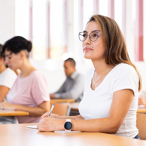 Eine junge Teilnehmerin sitzt an einem Tisch und schaut nach vorne. Sie hält einen Stift. An weiteren Tischen sitzen weitere Teilnehmer, diese sind unscharf.