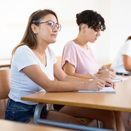 Zei Teilnehmde sitzen in einem Seminarraum. Im Fokus ist eine junge Frau mit Brille, die einer Person au&szlig;erhalb des Bildes zuh&ouml;rt. Sie hat einen Kugelschreiber in der Hand und ein Notizbuch vor sich liegen. Die anderen Frau hat ebenfalls Schreibger&auml;t und Hefte vor sich liegen.