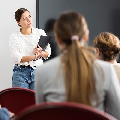 Eine junge Frau steht mit einer Mappe vor Publikum und spricht. Alle h&ouml;ren ihr aufmerksam zu. Im Hintergrund ist ein schwarzer Bildschirm.