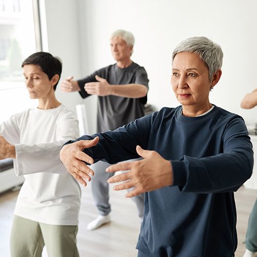 Zwei Frauen und ein Mann stehen in bequemer Kleidung in einem Raum und halten Arme und H&auml;nde vor sich ausgebreitet, als w&uuml;rden sie bestimmte Bewegungsabl&auml;ufe einstudieren.