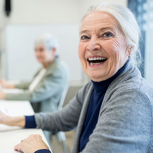 Eine &auml;ltere Frau sitzt in einem Seminarraum und lacht zur Seite in die Kamera. Im Hintergrund sind weitere &auml;ltere Personen unscharf zu erkennen.